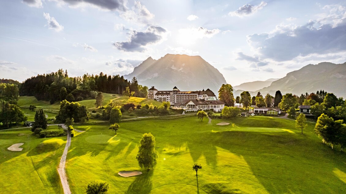 Eine weitläufige Golfanlage mit gepflegten grünen Rasenflächen und sanften Hügeln. Im Hintergrund thront ein majestätischer Berg unter einem bewölkten Himmel. | © Tom Sattler 