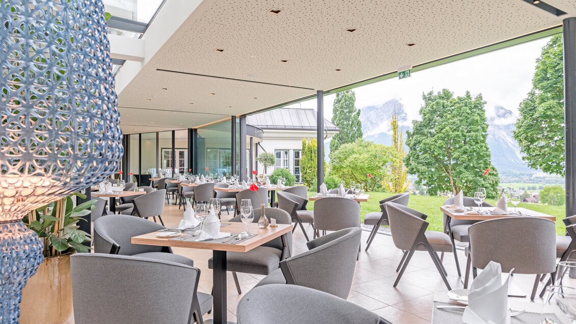 Ein modernes Restaurant mit eleganten Tischen und Stühlen. Im Hintergrund sind grüne Bäume und eine Berglandschaft sichtbar. | © Thomas Sattler
