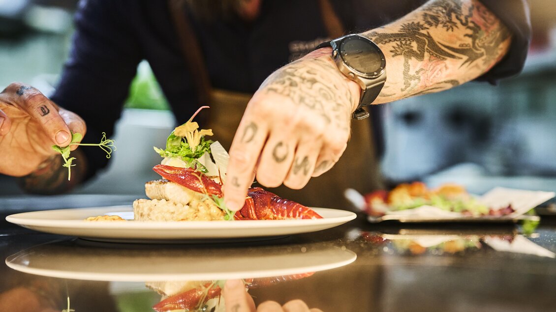 Ein Koch dekoriert ein Gericht auf einem weißen Teller. Es handelt sich um eine kunstvolle Anrichtung von Speisen mit frischen Zutaten. | © Thomas Sattler