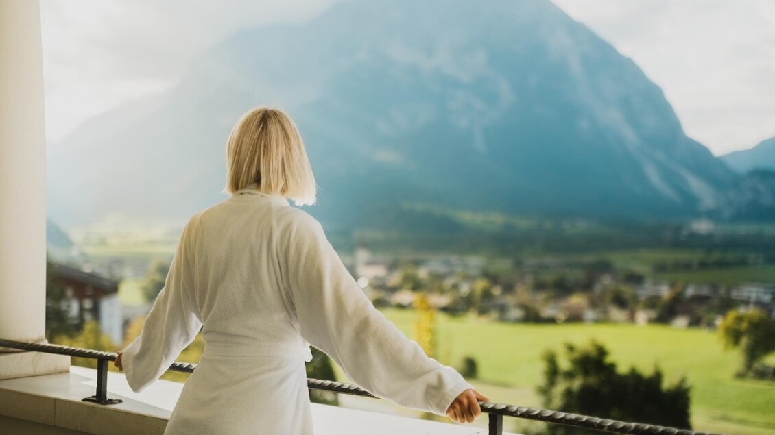 Eine Person in einem Bademantel steht auf einem Balkon und blickt auf eine beeindruckende Berglandschaft. Im Hintergrund sind grüne Wiesen und ein klarer Himmel zu sehen. | © Richard Schabetsberger