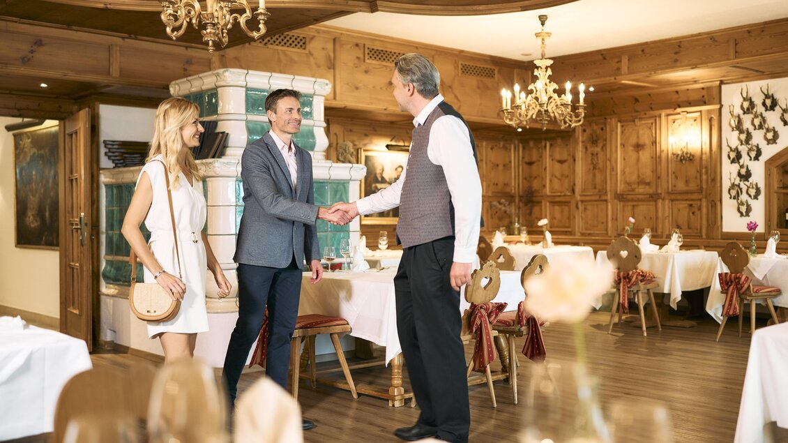 Ein elegantes Restaurant mit Holzdekor und stilvollen Tischen. Ein Gast wird herzlich von einem Mitarbeiter begrüßt, während eine Begleitperson daneben steht. | © Thomas Sattler