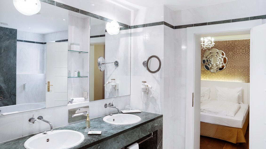 Ein modernes Badezimmer mit zwei Waschbecken und eleganten Spiegeln. Im Hintergrund ist das Schlafzimmer mit einem gemütlichen Bett sichtbar. | © Richard Schabetsberger
