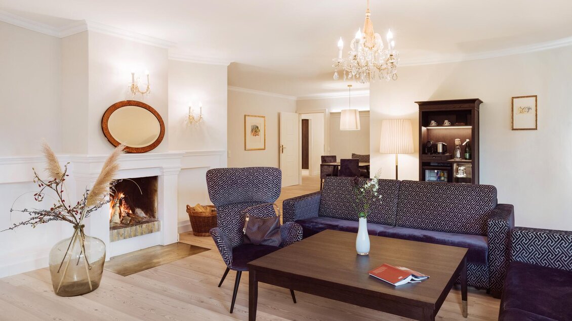 Ein stilvolles Wohnzimmer mit einem eleganten Kronleuchter und einer gemütlichen Sitzecke. Ein Kamin sorgt für eine einladende Atmosphäre, während dekorative Elemente den Raum verschönern. | © Richard Schabetsberger
