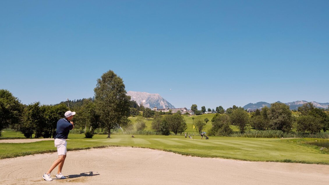 Ein Golfer schlägt einen Ball aus dem Bunker auf einem malerischen Golfplatz. Die Landschaft ist grün mit Bergen im Hintergrund und strahlend blauem Himmel. | © Edy Gröger