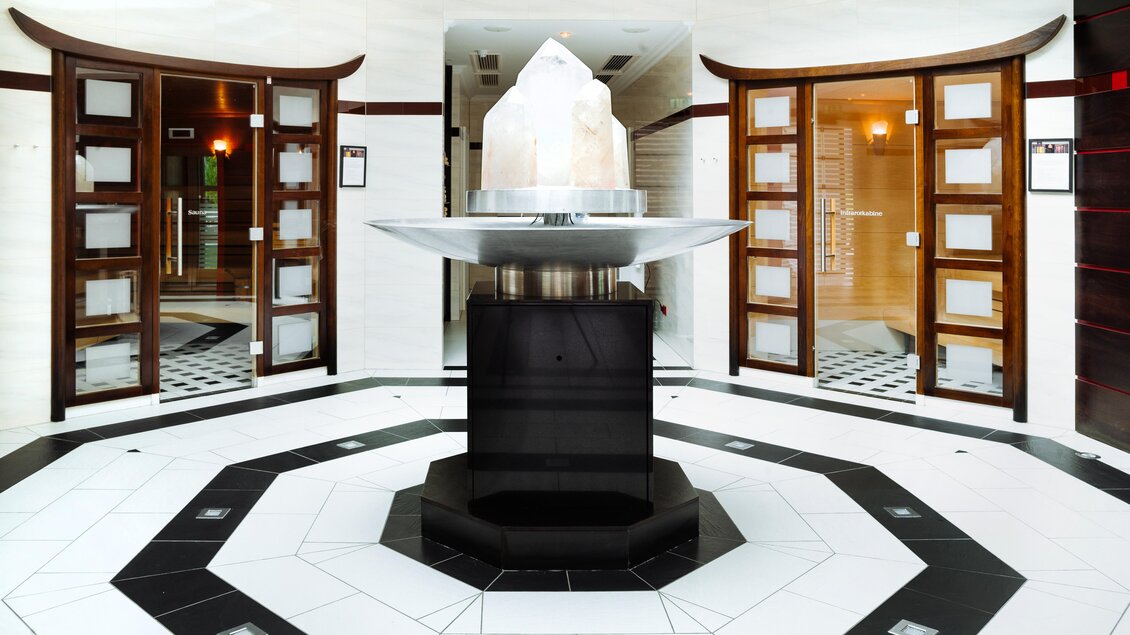 Ein eleganter Wellnessbereich mit einem zentralen Wasserspiel und stilvollen Holztüren. Der Boden ist in einem modernen Schwarz-Weiß-Design gestaltet. | © Richard Schabetsberger
