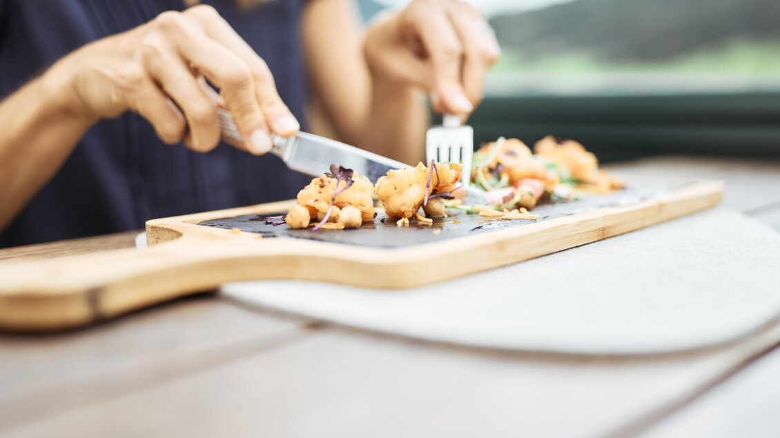 Eine Person schneidet und genießt ein Gericht auf einem Holzbrett. Frische Zutaten und eine schöne Präsentation sind sichtbar. | © Thomas Sattler
