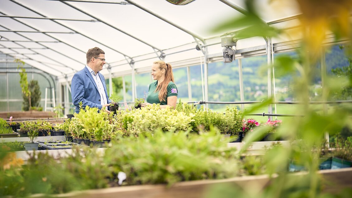 Ein Mann und eine Frau sprechen in einem Gewächshaus umgeben von Pflanzen. Die Atmosphäre ist freundlich und geschäftlich. | © Thomas Sattler