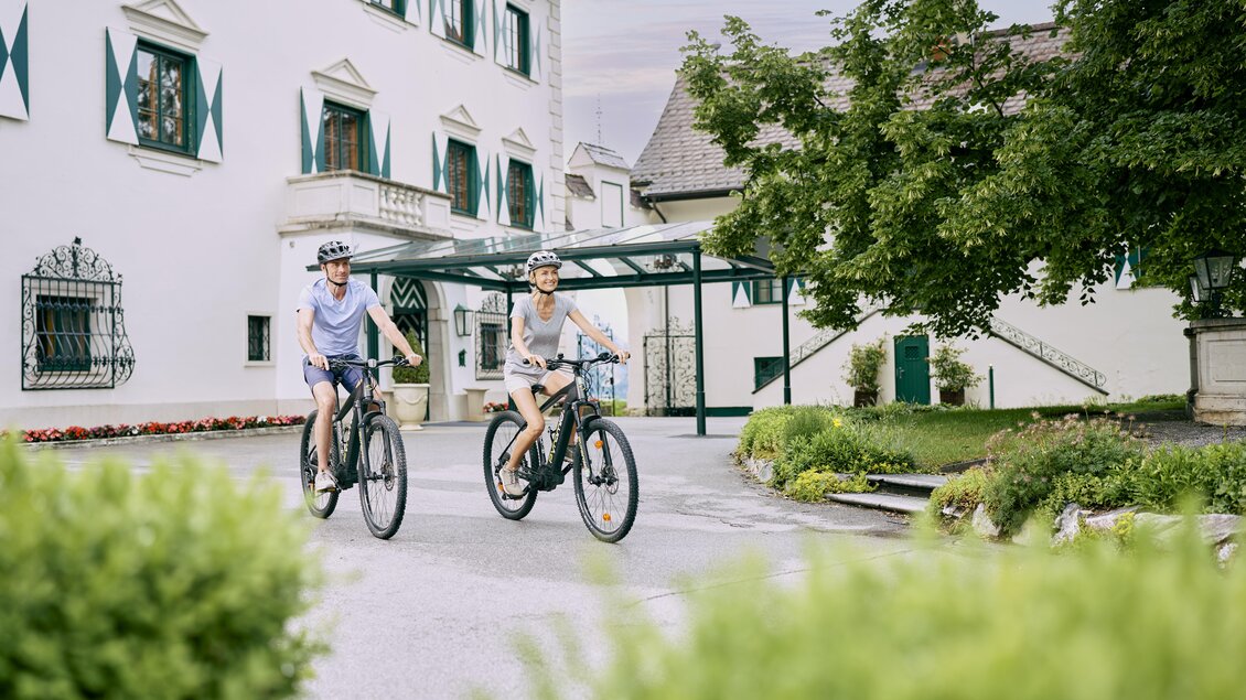 Zwei Personen fahren auf Fahrrädern in einem malerischen Innenhof. Im Hintergrund ist ein schönes Gebäude mit grünen Fenstern und ein gepflegter Garten zu sehen. | © Thomas Sattler