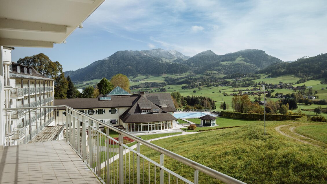 Eine ruhige Aussicht von einem Balkon auf eine grüne Landschaft mit Bergen im Hintergrund. Im Vordergrund sind Gebäude und ein Pool zu sehen. | © Richard Schabetsberger