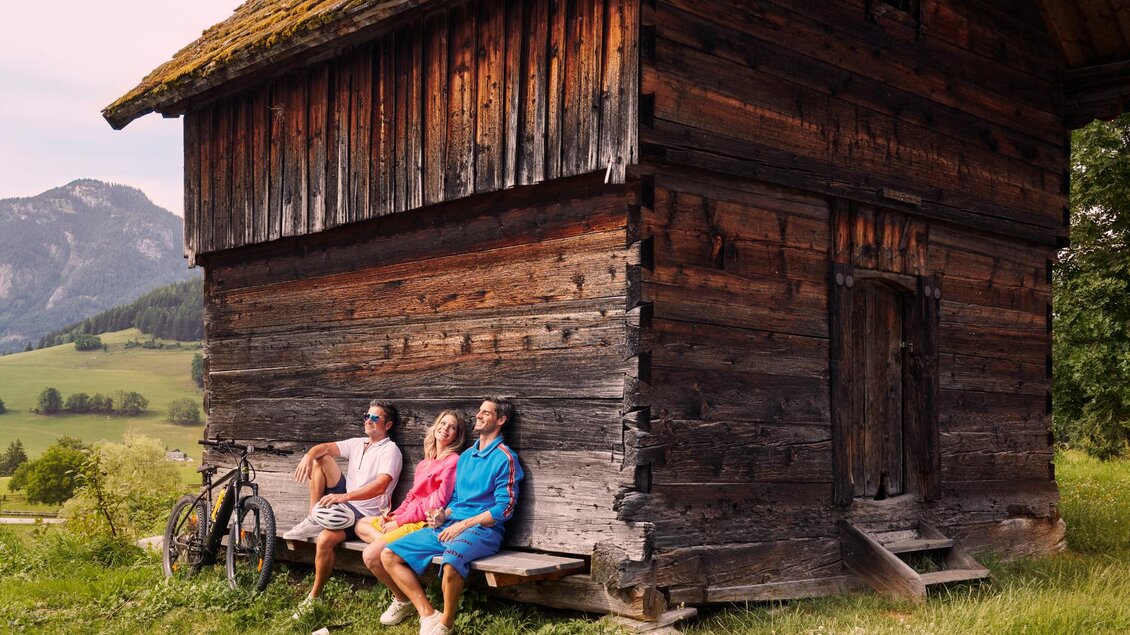Eine Gruppe von drei Freunden sitzt entspannt an einer rustikalen Holzhütte. Im Hintergrund sind sanfte Hügel und Berge sichtbar. | © Art in action