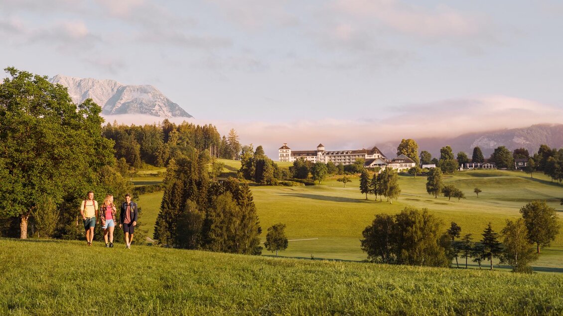 Eine Gruppe von Personen spaziert durch eine grüne Landschaft mit Bergen im Hintergrund. Im Hintergrund ist ein elegantes Gebäude sichtbar. | © Art in action