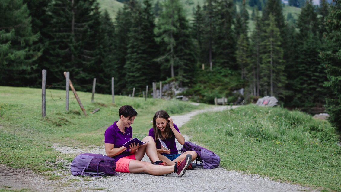 Zwei Personen sitzen auf einem Waldweg und schauen auf ein Tablet. Sie haben Rucksäcke dabei und umgeben von grünen Wiesen und Bäumen. | © Indrich Fotografie 