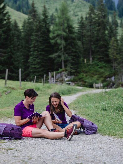 Two people are sitting on a forest path and looking at a tablet. They have backpacks with them and are surrounded by green meadows and trees. | © Indrich Fotografie 