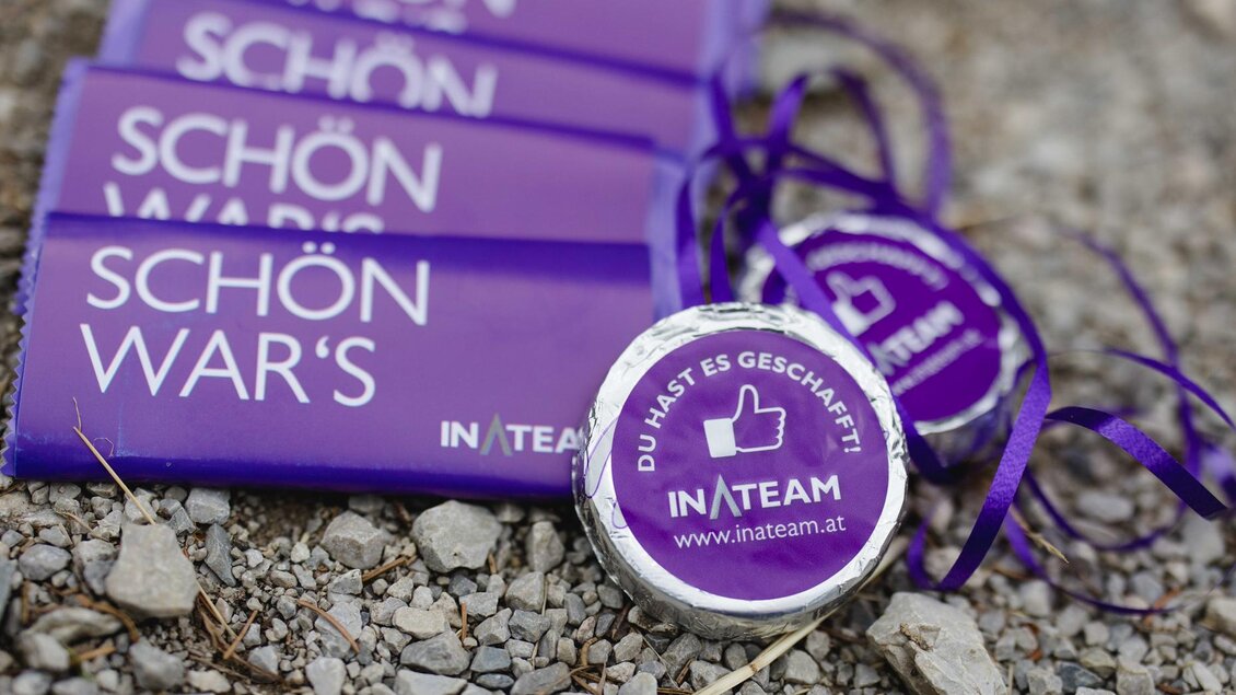 Eine Gruppe von lila Bändern und Medaillen mit den Aufschriften "SCHÖN WAR'S" und "DU HAST ES GESCHAFFT!" liegt auf einem Kiesboden. Die Medaillen gehören zum INATEAM und feiern Erfolge oder Teilnahmen. | © Indirch Fotografie