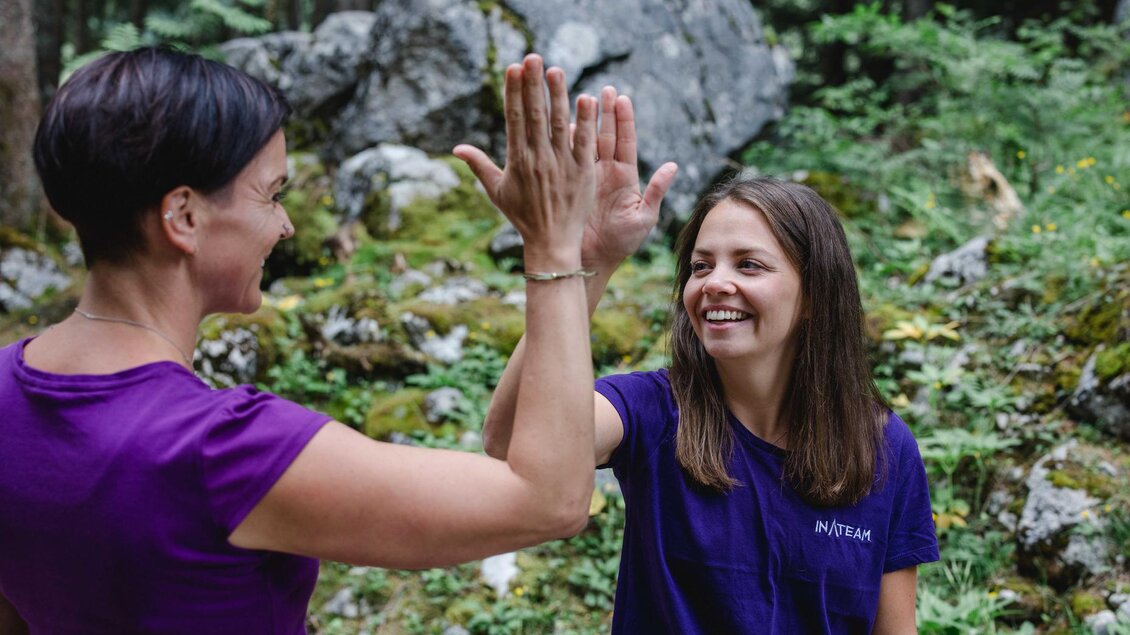 Zwei Frauen geben sich ein High Five in einer grünen, bewaldeten Umgebung. Beide tragen lila T-Shirts und lachen fröhlich. | © Indirch Fotografie