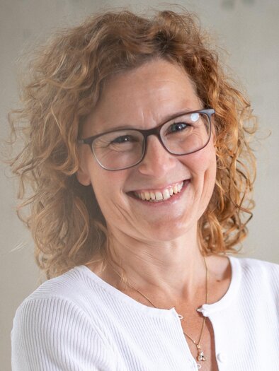 A smiling woman with curly hair is wearing glasses. She has a friendly and open personality.