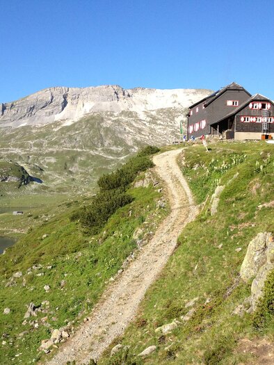 Ein malerisches Bergland mit einer Wiese und einem klaren See. Im Hintergrund sind majestätische Berge und eine Hütte zu sehen. | © Gerhard Pilz