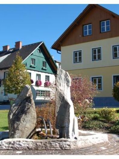A beautiful stone fountain in a garden. In the background, several colorful houses can be seen. | © TVB Gröbmingerland
