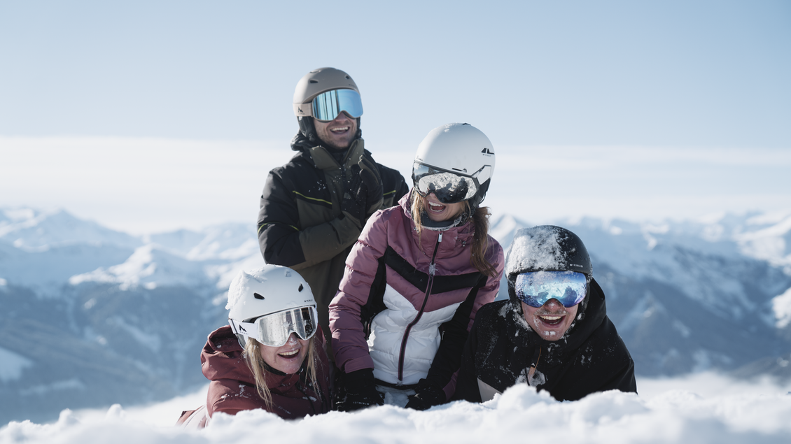 lächelende junge Menschen im Skianzug und Skihelm sowie Skibrille die im Schnee spielen bei schönem Wetter. | © Intersport Gerhardter