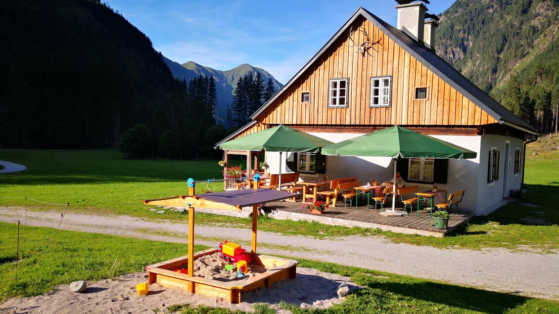 Ein gemütliches Holzhaus mit grünen Sonnenschirmen steht in einer malerischen Landschaft. Vor dem Haus befindet sich ein Sandkasten für Kinder. | © Familie Moitzi