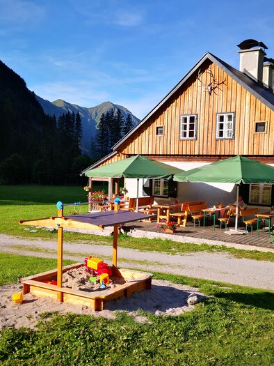 A cozy wooden house with green umbrellas is situated in a picturesque landscape. In front of the house, there is a sandbox for children. | © Familie Moitzi