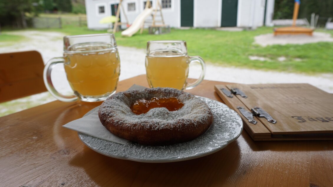 Ein leckerer Krapfen mit Puderzucker und einer Füllung aus Marmelade liegt auf einem Teller. Dazu stehen zwei Gläser mit einem hellen Getränk auf dem Tisch, im Hintergrund ist ein Gebäude zu sehen. | © Familie Moitzi
