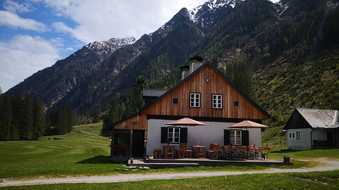 Ein gemütliches Holzhaus in den Bergen mit schneebedeckten Gipfeln im Hintergrund. Vor dem Haus stehen Tische und Stühle auf einer grünen Wiese. | © Familie Moitzi