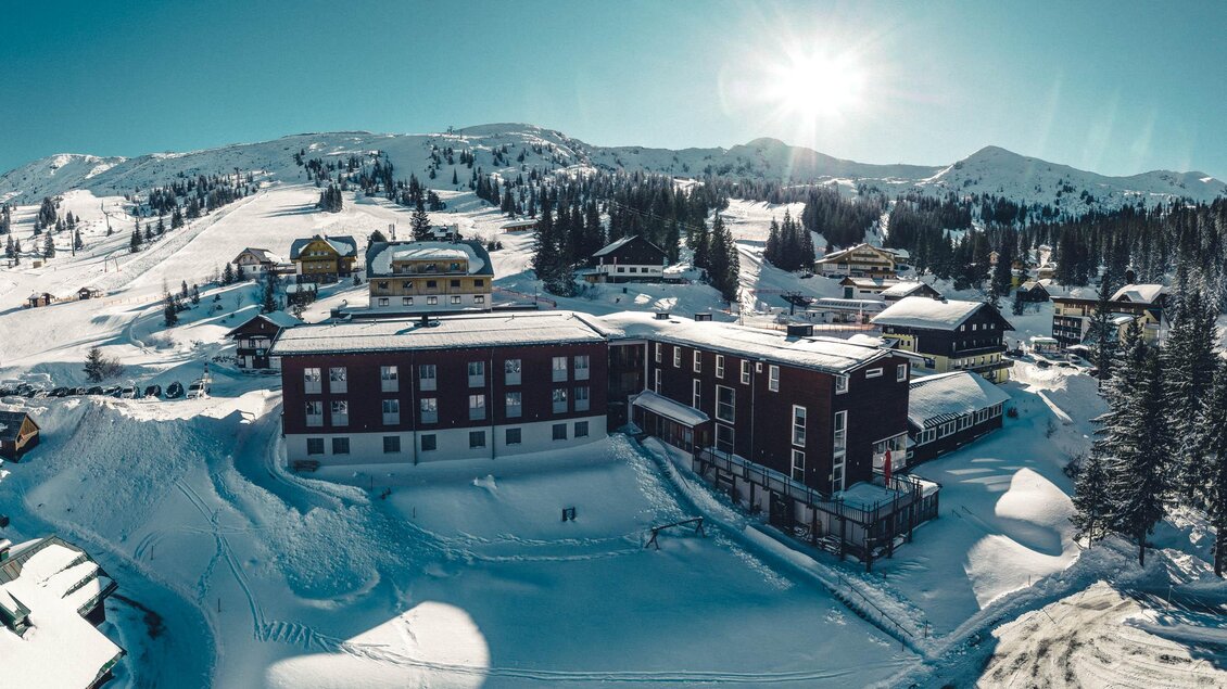 Eine verschneite Berglandschaft mit einem Hotel und einigen kleinen Chalets. Die Sonne scheint über die Berge und die umliegende Natur.