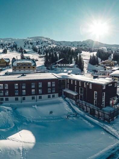 A snowy mountain landscape with a hotel and some small chalets. The sun shines over the mountains and the surrounding nature.