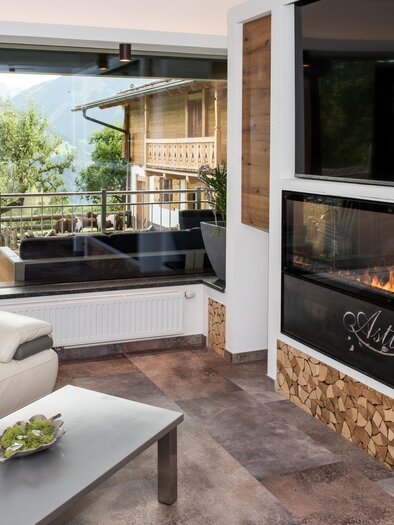 Ein modernes Wohnzimmer mit einem großen, offenen Kamin und einer gemütlichen Couch. Hinter dem Fenster sieht man eine schöne Aussicht auf die Berge und die Natur. | © Shooting Star