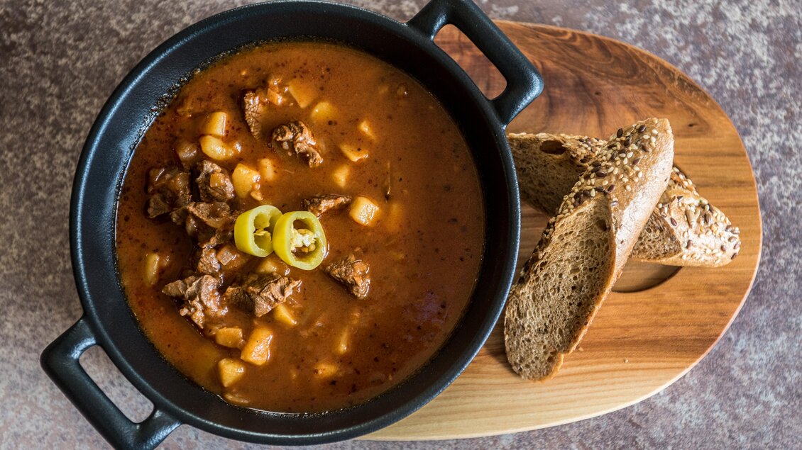 Eine leckere Suppe mit Fleisch, Kartoffeln und Paprika, serviert in einem schwarzen Topf. Dazu gibt es frisches Vollkornbrot auf einem Holzbrett. | © Shooting Star