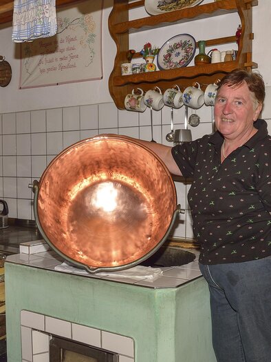 Eine Person steht in einer gemütlichen Küche und hält einen großen Kupfertopf. Im Hintergrund sind Holzscheite und Küchengeräte zu sehen. | © Herfried Marek