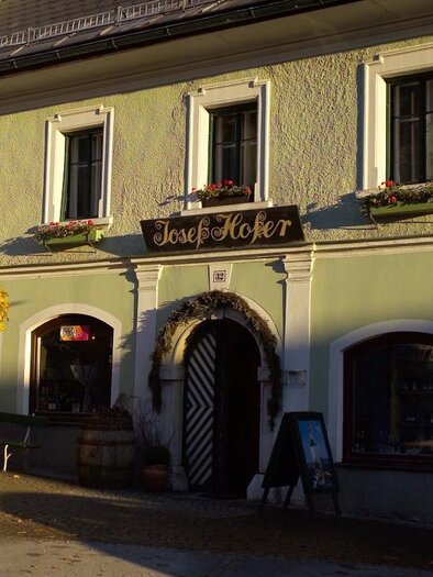 A charming building with a green facade and blooming windows. Below is a sign with the name "Josef Hofer". | © Josef Hofer e.U.