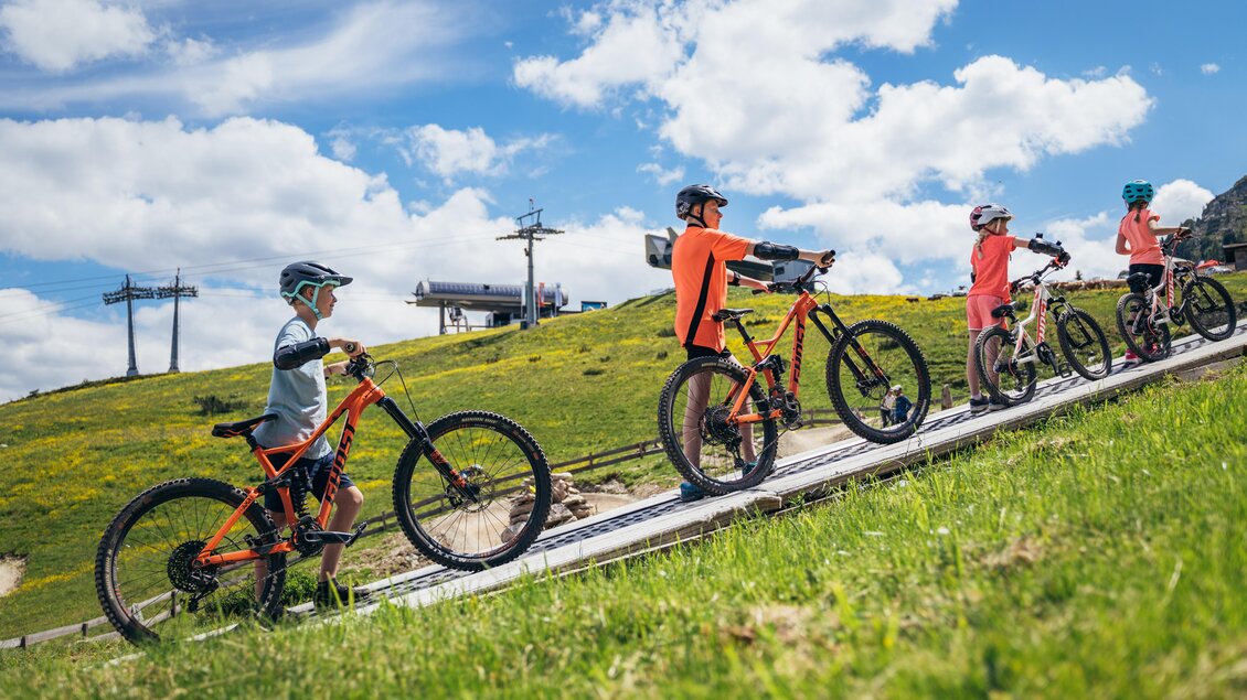 Eine Gruppe von Kindern fährt mit Mountainbikes einen Hang hinauf. Der Himmel ist blau mit weißen Wolken und im Hintergrund sind Skilifte zu sehen. | © Gerald Grünwald