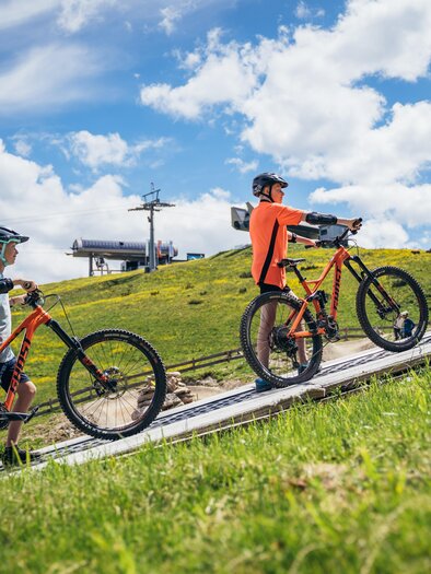 A group of children is riding mountain bikes up a hill. The sky is blue with white clouds, and in the background, ski lifts can be seen. | © Gerald Grünwald