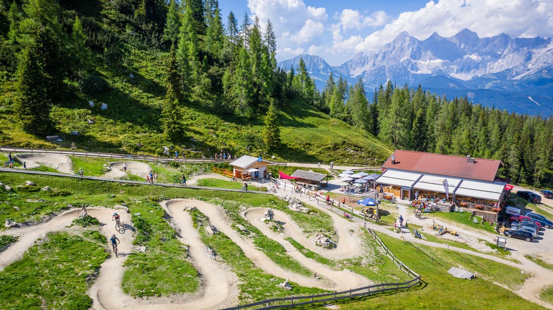 Eine malerische Berglandschaft mit einem kurvenreichen Parcours für Bikes. Im Hintergrund sieht man eine Hütte und majestätische Berge unter einem klaren Himmel. | © Christoph Oberschneider