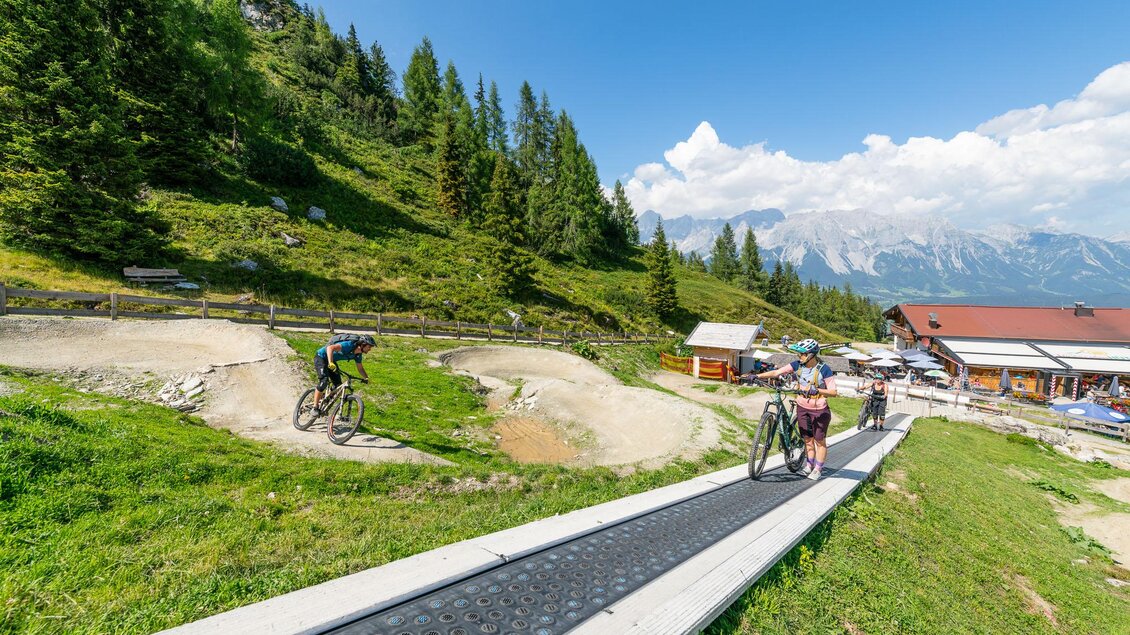 Ein sonniger Tag in den Bergen mit Radfahrern auf einem Fahrradparcours. Im Hintergrund sind grüne Wiesen und Berge zu sehen. | © Christoph Oberschneider
