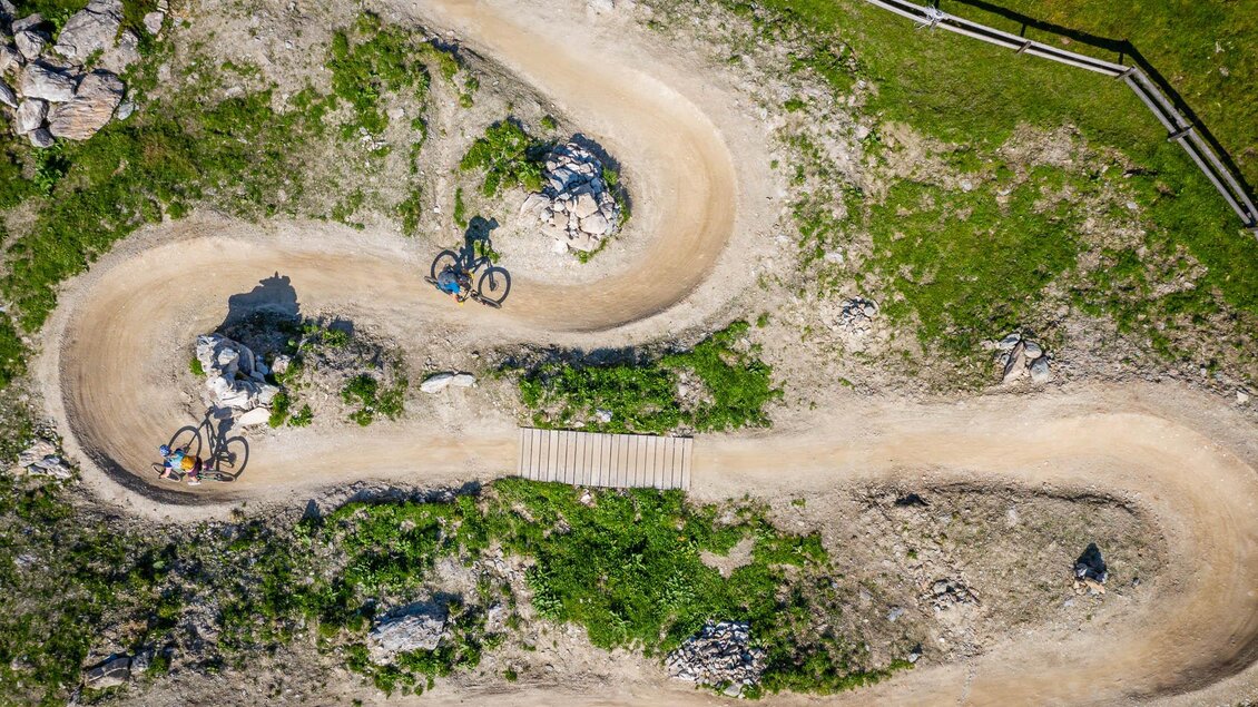 Eine kurvenreiche Mountainbike-Strecke umgeben von grünem Gras und Felsen. Zwei Radfahrer fahren auf dem Trail. | © Christoph Oberschneider