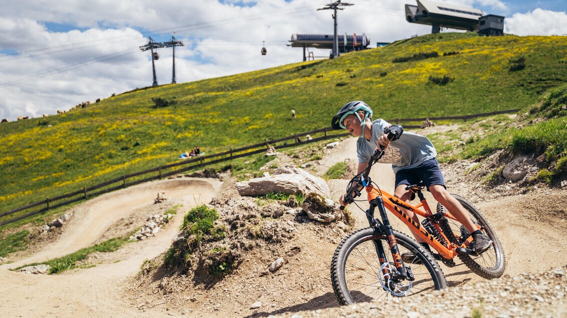 Ein Junge fährt mit dem Mountainbike auf einem abfallenden Trail in einer grünen Landschaft. Im Hintergrund sind eine Seilbahn und eine blühende Wiese zu sehen. | © Gerald Grünwald