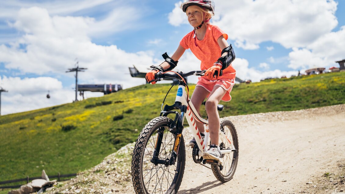 Ein Mädchen fährt mit einem Mountainbike auf einem Schotterweg. Es ist ein schöner, sonniger Tag mit grünen Wiesen im Hintergrund. | © Gerald Grünwald