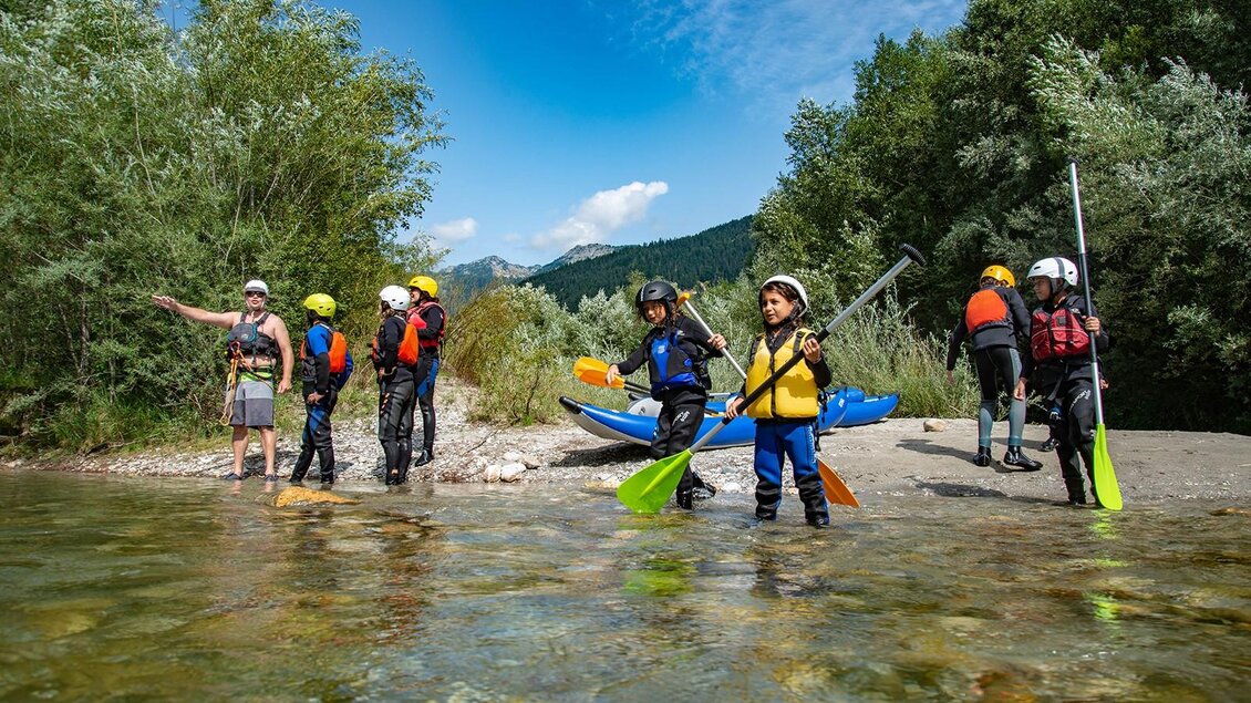 Eine Gruppe von Kindern steht am Ufer eines Flusses, bereit zum Kajakfahren. Es gibt Bäume im Hintergrund und eine sonnige, wolkenlose Himmel. | © Kajaktiv