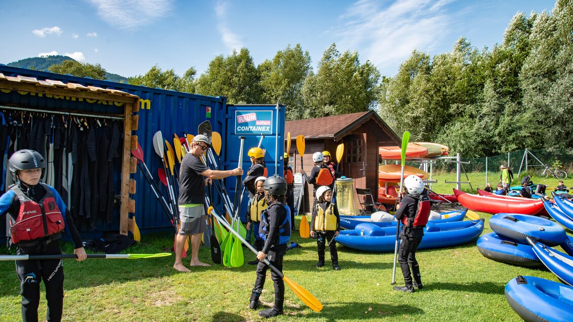 Eine Gruppe von Kindern bereitet sich auf das Kajakfahren vor. Sie stehen auf einer Wiese neben Kajaks und Paddeln, während ein Trainer ihnen Anweisungen gibt. | © Kajaktiv