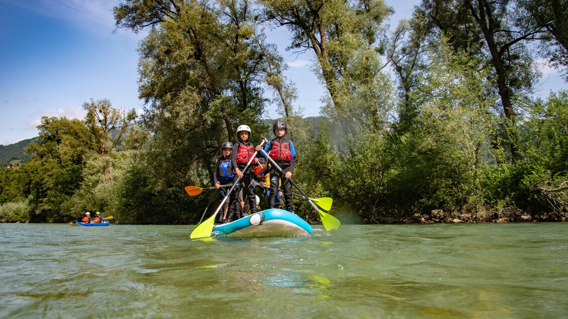 Eine Gruppe von vier Personen steht auf einem Stand-Up-Paddle-Board im Wasser. Umgeben von grünen Bäumen und einer ruhigen Landschaft genießen sie einen sonnigen Tag. | © Kajaktiv