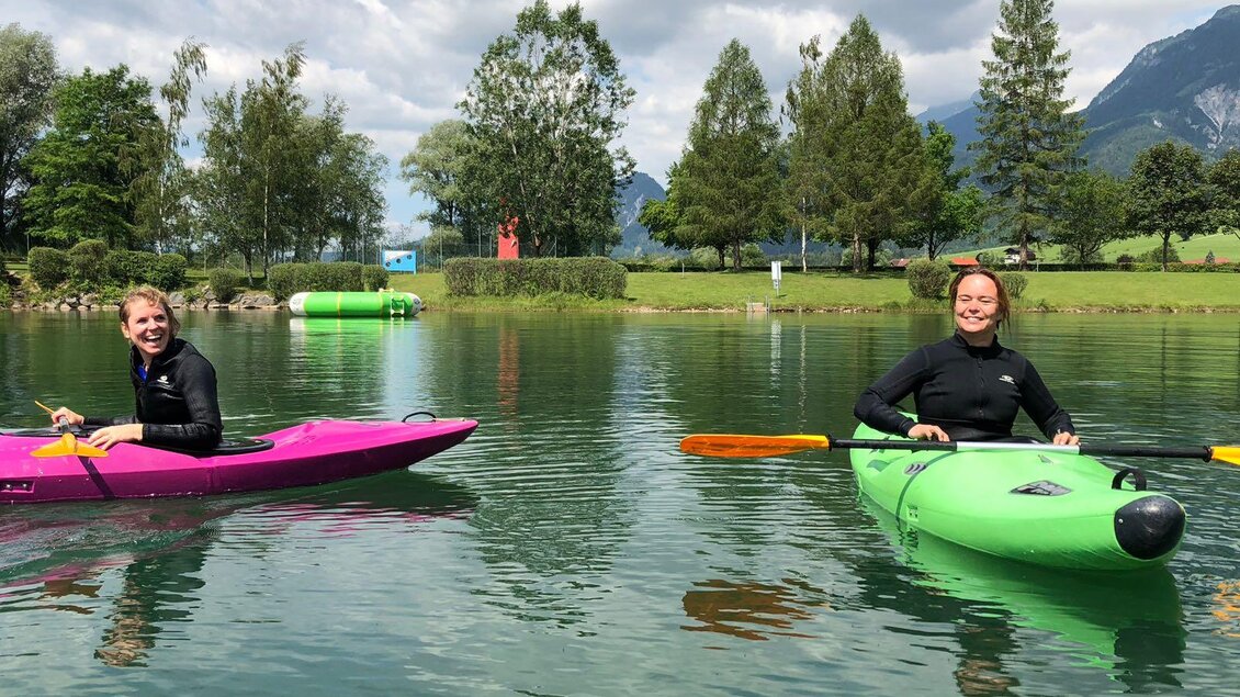 Zwei Personen paddeln fröhlich mit Kajaks auf einem ruhigen See. Im Hintergrund sind grüne Bäume und die Berge zu sehen. | © Kajaktiv