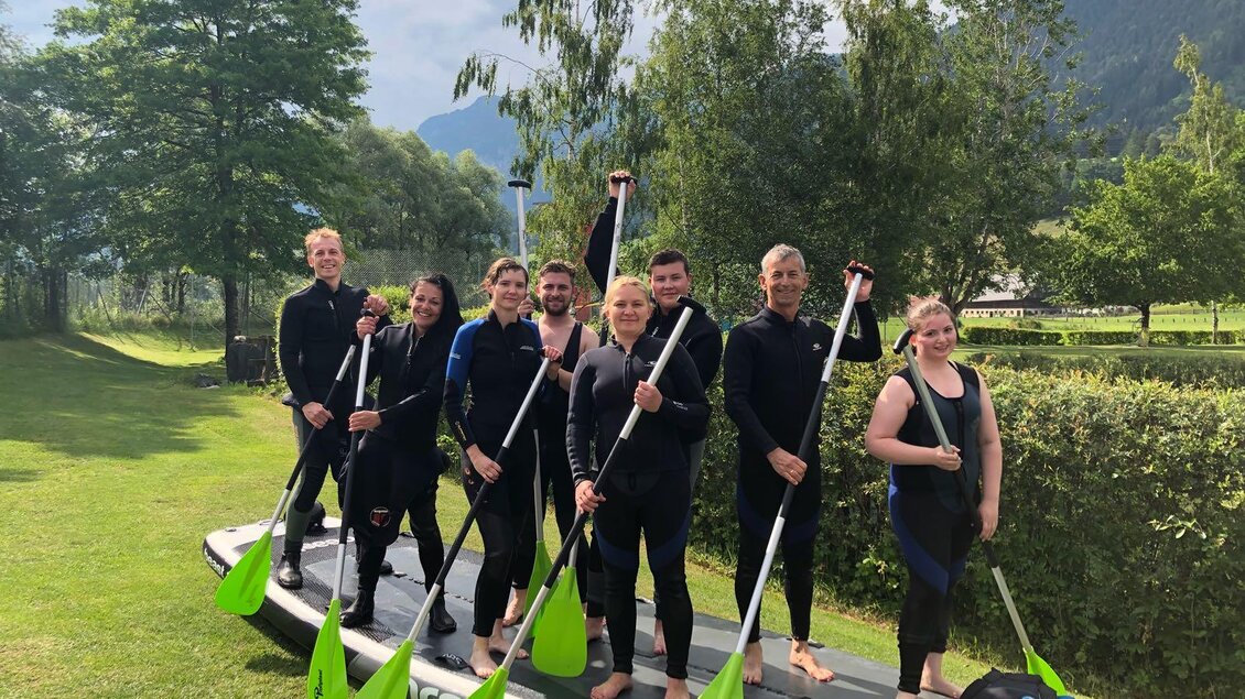 Eine Gruppe von acht Personen steht auf einem Stand-Up-Paddle-Board. Sie tragen Neoprenanzüge und halten Paddel in der Hand, umgeben von grüner Landschaft und Bäumen. | © Kajaktiv