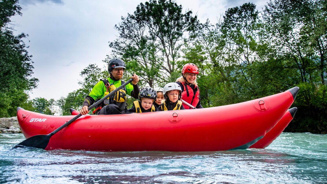 Eine Familie mit zwei Kindern paddelt in einem roten Kajak auf einem klaren Fluss. Im Hintergrund sind grüne Bäume und ein bewölkter Himmel sichtbar. | © Kajaktiv