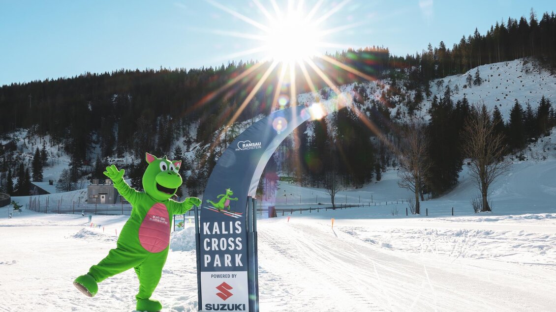 Ein grünes Maskottchen steht vor einem Banner des Kalis Cross Parks im Schnee. Die Sonne scheint hell über der winterlichen Landschaft. | © Loretta Kvitek
