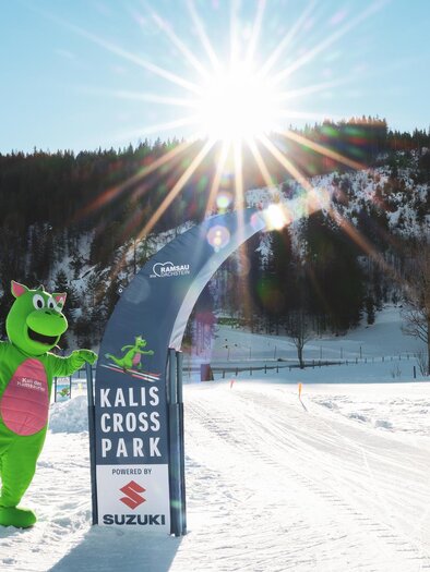 A green mascot stands in front of a banner of the Kalis Cross Park in the snow. The sun shines brightly over the wintry landscape. | © Loretta Kvitek
