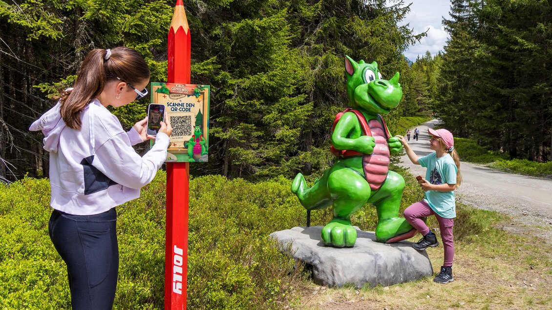 Ein Mädchen fotografiert eine grüne Drachenfigur, während ein Junge davor steht. Im Hintergrund sind grüne Bäume und ein Waldweg zu sehen. | © Rittisberg