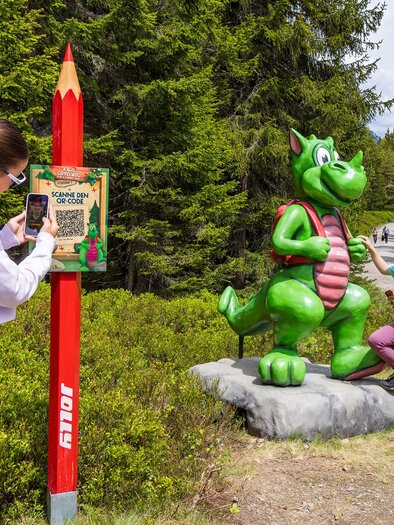 A girl is photographing a green dragon figure while a boy stands in front of it. In the background, green trees and a forest path can be seen. | © Rittisberg